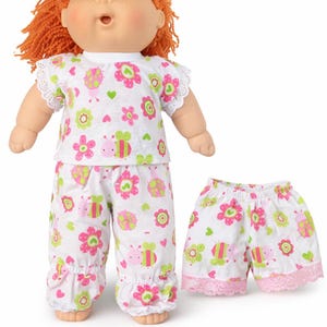 May include: A doll with orange yarn hair and green eyes wearing a white short-sleeved top and long pants featuring a pink and green floral and insect pattern. Beside the doll are matching white shorts with a pink lace trim and the same pattern.