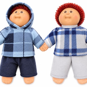 May include: Two dolls with round faces, tan skin, and red yarn hair. The left doll wears a blue plaid hooded jacket and dark blue shorts. The right doll wears a blue and white plaid long-sleeve shirt and light grey trousers. Both dolls are holding hands.