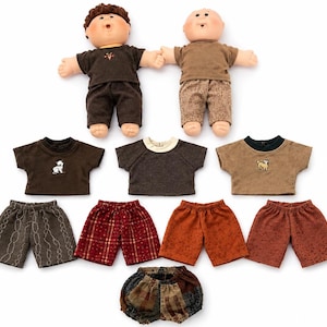 May include: Two dolls with light skin tones, one with brown hair and one bald, wearing brown shirts and trousers. Below them are three short-sleeved shirts in brown and grey, four pairs of shorts in various patterns and colours, and one pair of patterned bloomers.