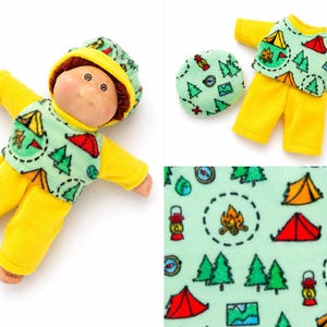May include: A doll with brown hair wearing a yellow and light green outfit featuring a camping-themed print of tents, trees, lanterns, and compasses. The set includes a matching hat and a separate shirt and pants outfit with the same pattern.