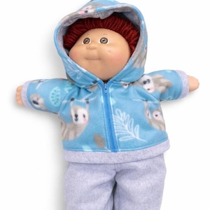 May include: A Cabbage Patch Kid doll dressed in a blue fleece zip-up hoodie and grey sweatpants. The hoodie has a sloth pattern and a matching hood. The doll has red yarn hair and a smiling expression.