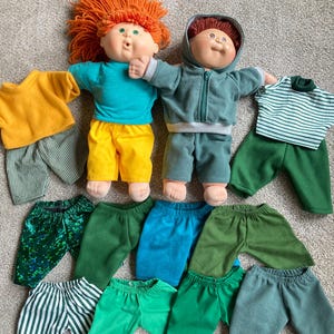 May include: Two Cabbage Patch Kids dolls with various outfits. One doll has orange hair and is wearing a teal shirt and yellow shorts. The other doll has brown hair and is wearing a green hooded jacket and pants. Several pairs of pants and shirts are also displayed.