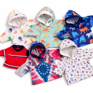 May include: A collection of colorful hooded sweatshirts for children. The sweatshirts feature various patterns, including foxes, tigers, and floral designs. One sweatshirt has a red, white, and blue tie-dye pattern with stars. The hoodies are made of soft fabric.