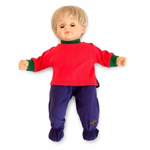 May include: A doll with blonde hair and a fair complexion wearing a long-sleeved red shirt with a green mock neck and green cuffs, paired with purple trousers that have gathered ankles and a small embroidered graphic on the lower left leg.
