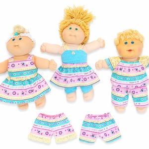 May include: Three Cabbage Patch Kid dolls in colourful outfits. The dolls have blonde hair and blue eyes. The outfits include dresses and shorts with a pink, blue, and yellow butterfly pattern. Two pairs of matching shorts are also included.