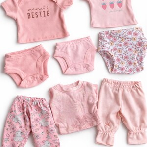 May include: A collection of doll clothing in shades of pink, including two t-shirts with prints, three pairs of briefs, one pair of patterned pants with rabbits, one sleeveless top, and one pair of solid pink pants with a small decorative detail.