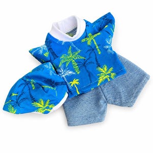 May include: A blue doll's outfit comprising a short-sleeved shirt, shorts, and a visor. The shirt and visor display a palm tree design in yellow, green, and white on a blue backdrop. The shorts are a solid blue denim.