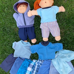 May include: Two Cabbage Patch Kids dolls with red and brown hair, wearing blue and purple outfits. Additional doll clothes in various shades of blue are arranged on the grass, including shirts and shorts. The dolls are lying on a bed of green grass.
