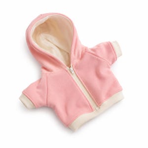 May include: A light pink zip-up hoodie featuring a cream-colored interior lining, cream-colored sleeve cuffs, and a cream-colored bottom hem. The garment includes a central white zipper that extends from the neckline to the bottom hem.