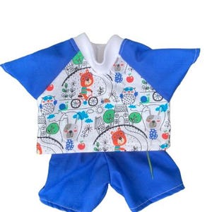 May include: A baby's two-piece outfit. The top is white with a colourful cartoon print and blue sleeves, and the shorts are blue. The top has a crew neck and the print includes bears, owls, houses, and bicycles.