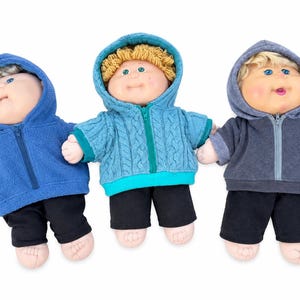 May include: Three dolls with soft vinyl faces and yarn hair, each wearing a hooded zip-up jacket and black trousers. The left doll wears a blue fleece jacket, the centre doll a teal cable-knit jacket, and the right doll a charcoal grey hoodie.