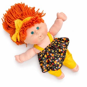 May include: A doll with bright orange yarn hair, a yellow hair bow, and blue eyes. The doll has freckles and wears a black dress featuring a colorful candy corn pattern over yellow pants. The doll is positioned with arms outstretched.