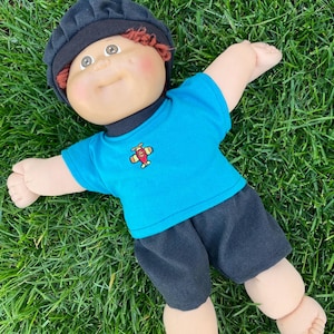 May include: A Cabbage Patch Kid doll, lying on green grass, wearing a black cap, a blue t-shirt with a red and yellow airplane graphic, and black shorts. The doll has red hair and rosy cheeks. The doll's arms and legs are outstretched.