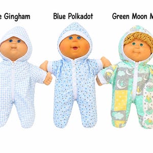 May include: Three dolls with round faces and blue eyes, each wearing a hooded zip-up sleeper. The first is labeled Blue Gingham, the second Blue Polkadot, and the third Green Moon Monkey, featuring a pattern of moons, clouds, and monkeys.
