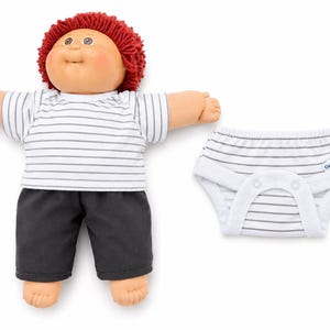 May include: A doll with red yarn hair, a round face, and rosy cheeks, wearing a white t-shirt with thin horizontal grey stripes and solid black shorts. Beside the doll is a white nappy with thin grey stripes and a small Gerber brand tag.