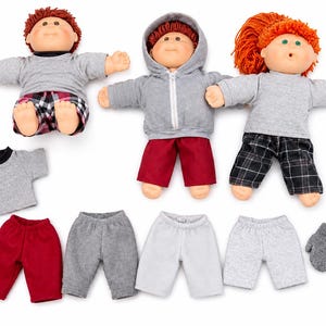 May include: Three dolls with yarn hair and plastic faces, dressed in gray shirts, hoodies, and plaid or solid pants. The set includes extra clothing items: a gray t-shirt, four pairs of pants in red, gray, white, and light gray, and gray mittens.