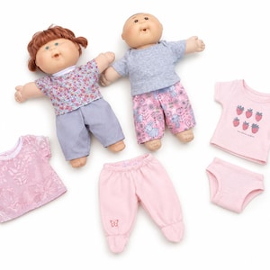 May include: Two dolls with round faces and yarn hair, one with reddish-brown hair and one with short brown hair. They wear floral and patterned tops with solid and printed trousers. Included are a pink textured top, pink trousers, and a pink strawberry-print shirt.