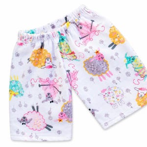 May include: White shorts featuring a colorful all-over print of stylized sheep in shades of pink, yellow, blue, and grey. The sheep are depicted with various expressions and accessories, interspersed with small grey line-drawn flower outlines.