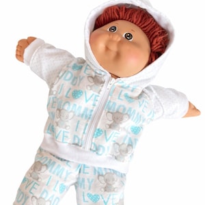 May include: A doll with reddish-brown yarn hair wearing a white hooded zip-up jacket and matching trousers. The clothing features a repeating pattern of light blue hearts, grey elephants, and the text I LOVE MOMMY and I LOVE DADDY in blue.