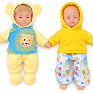 Peut inclure: Deux poupées bébé se tenant la main. La poupée de gauche a des cheveux blonds, un sweat à capuche jaune avec des oreilles d'ours et un haut bleu avec un lion. La poupée de droite est chauve, porte un sweat jaune et un pantalon à motifs animaux.