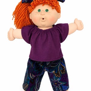 May include: A doll with bright orange yarn hair styled in a ponytail with a dark blue patterned headband. The doll has green eyes, freckles, and a small open mouth. It wears a solid purple short-sleeved top and dark blue corduroy trousers.