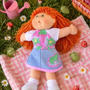 May include: A vintage doll with orange yarn hair, green eyes, and freckles, wearing a white-sleeved top and a pink frog-print bodice, a blue denim skirt, and white shoes. The doll is lying on a pink gingham blanket, surrounded by daisies, toy mushrooms, and green frog figurines.