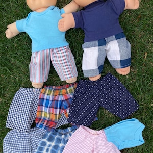 May include: Two Cabbage Patch Kids dolls wearing blue and navy shirts with various patterned shorts. The shorts include stripes, plaid, polka dots, and gingham. A light blue onesie is also included.