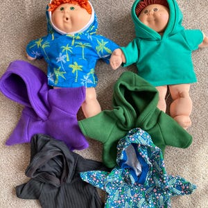 May include: Several Cabbage Patch Kids dolls wearing colorful hoodies. The hoodies are in various colors and patterns, including blue with palm trees, solid green, purple, black, and a floral print. The dolls have orange and brown hair.