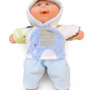 May include: A Cabbage Patch Kids doll wearing a blue and white hooded outfit with an elephant design. The doll has blue eyes and a smiling expression. The doll is wearing light blue pants.