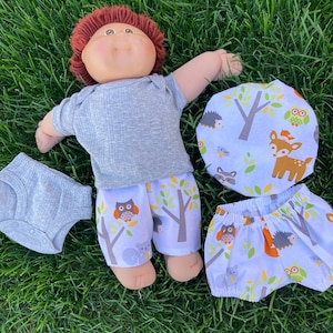 May include: A doll wearing a gray ribbed onesie, white shorts with an owl and tree print, and a matching hat. Additional gray diaper and shorts with the same print are also included. The doll has red hair and is lying on green grass.