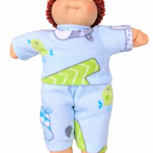 May include: A doll with a round face, rosy cheeks, and short brown yarn hair. The doll wears a light blue long-sleeved shirt and matching trousers featuring a repeating pattern of green geometric shapes, blue birds, and grey clouds on a soft fabric.