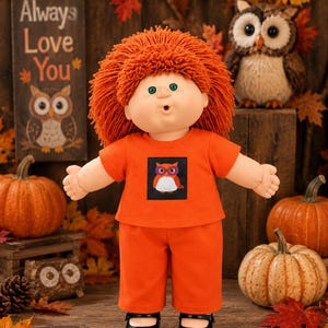 May include: A doll with bright orange yarn hair, green eyes, and freckles, wearing an orange short-sleeved shirt with a graphic of an owl wearing purple glasses and matching orange pants, paired with black shoes.