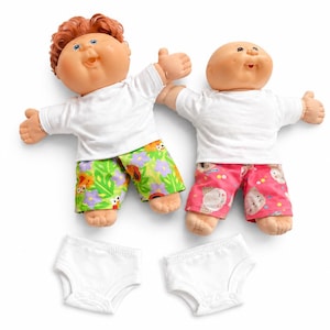 May include: Two dolls with round faces and soft bodies. One doll has reddish-brown hair, the other is bald. Both wear plain white t-shirts. The first doll wears green shorts with a floral pattern, the second wears pink shorts with a cat print. Two white briefs are placed below.