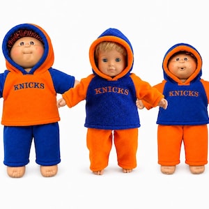 May include: Three dolls wearing hooded sweatshirts and pants in blue and orange. The sweatshirts feature the text KNICKS in blue or orange lettering. The dolls have different hair colors and styles, including brown yarn hair and blonde hair.