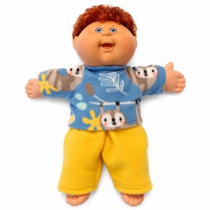 May include: A doll with a round face, blue eyes, and short curly reddish-brown hair. The doll wears a blue long-sleeved shirt featuring a pattern of small grey animals, white leaves, and yellow flowers, paired with solid bright yellow trousers.