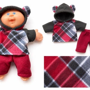 May include: A doll wearing a hooded fleece sweatshirt with a red, black, and white plaid pattern and solid black sleeves, paired with solid dark red fleece pants. The set includes a matching hooded sweatshirt and pants, plus a fabric swatch.