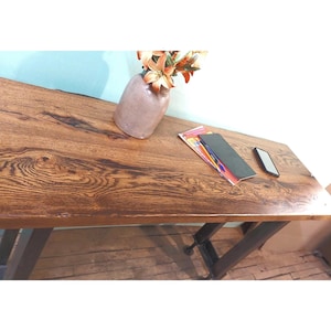 May include: A wooden desk with a dark brown, textured surface. The desk has a metal frame and is topped with a vase of flowers, a stack of papers, and a smartphone. The desk is suitable for a home office or workspace.