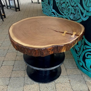 May include: A round, wooden side table with a natural edge and a light brown finish. The tabletop is approximately 30 inches in diameter and features a crack reinforced with two wooden butterfly joints. The base is made of black metal.