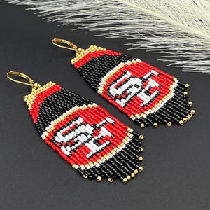 May include: A pair of beaded earrings featuring a red, black, white, and gold color scheme. The earrings display the letters "SF" in white, with a fringe at the bottom. The earrings have gold-colored ear wires.