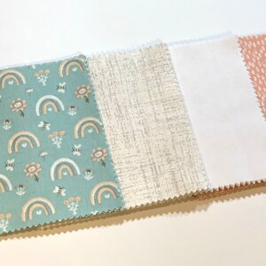 May include: A collection of fabric swatches with various patterns. One swatch features a teal background with rainbows, flowers, and bees. Other swatches include a textured gray, solid white, and a pink dotted design. The edges are serged.