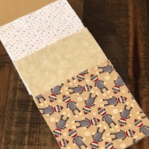 May include: Four fabric squares stacked on top of each other. The top square is a brown fabric with a white polka dot pattern. The second square is a white fabric with a brown and blue criss-cross pattern. The third square is a beige fabric with a subtle texture. The bottom square is a beige fabric with a repeating pattern of gray sock monkeys wearing red hats.