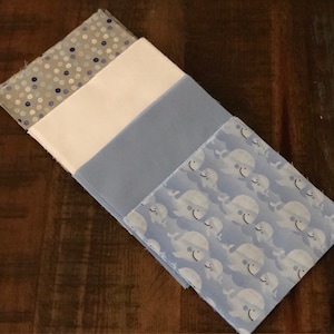 May include: Four fabric squares stacked on top of each other. The top square is a light gray with white polka dots. The second square is white. The third square is light blue. The bottom square is light blue with a white whale pattern.