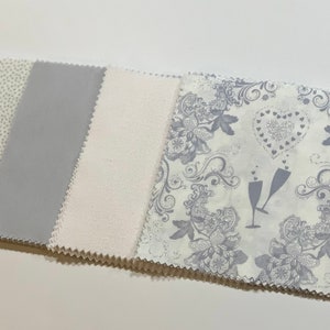 May include: Four fabric squares in a stack. The top square has a white background with a silver floral pattern and two silver champagne glasses. The second square is white with a subtle silver shimmer. The third square is a solid silver color. The bottom square has a white background with a silver polka dot pattern.
