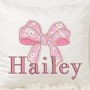 Könnte beinhalten: Ein weißes Kissen mit einer rosa floralen Stickerei eines Bogens und dem Namen "Hailey".