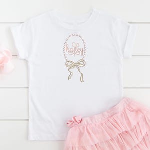 May include: White t-shirt with a pink pearl necklace and bow design, the name "hailey" is written in pink inside the necklace. The shirt is paired with a pink tulle skirt.