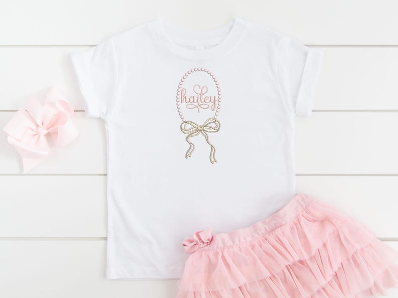 May include: White t-shirt with a pink pearl necklace and bow design, the name "hailey" is written in pink inside the necklace. The shirt is paired with a pink tulle skirt.