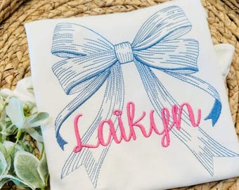 May include: A white t-shirt featuring an embroidered light blue bow design with detailed stitching. The name Laikyn is embroidered across the center of the bow in a bright pink cursive font, creating a clear and decorative graphic element.