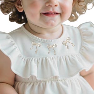 May include: A white baby dress with a ruffled neckline and three embroidered bows. The dress has short sleeves and a gathered skirt.