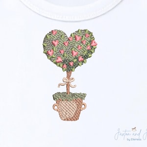 Könnte beinhalten: Gesticktes Design eines herzförmigen Topiaries in einem Weidenkorb. Der Baum hat rosa herzförmige Blüten und grüne Blätter. Eine beige Schleife ist um den Stamm gebunden. Das Design befindet sich auf weißem Hintergrund.