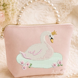 May include: A light pink pouch featuring an embroidered white swan wearing a gold crown, floating on a light green patch with small pink flowers. The bag has a gold zipper and a handle made of white faux pearls. The text Jaxton and Jill is visible.
